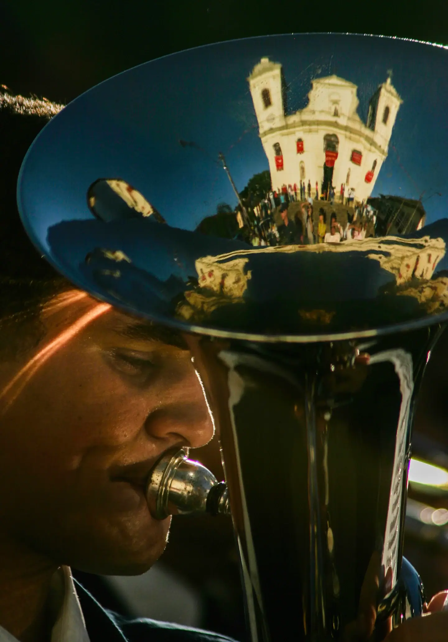 Músico toca tuba com reflexos da igreja e da multidão na campana