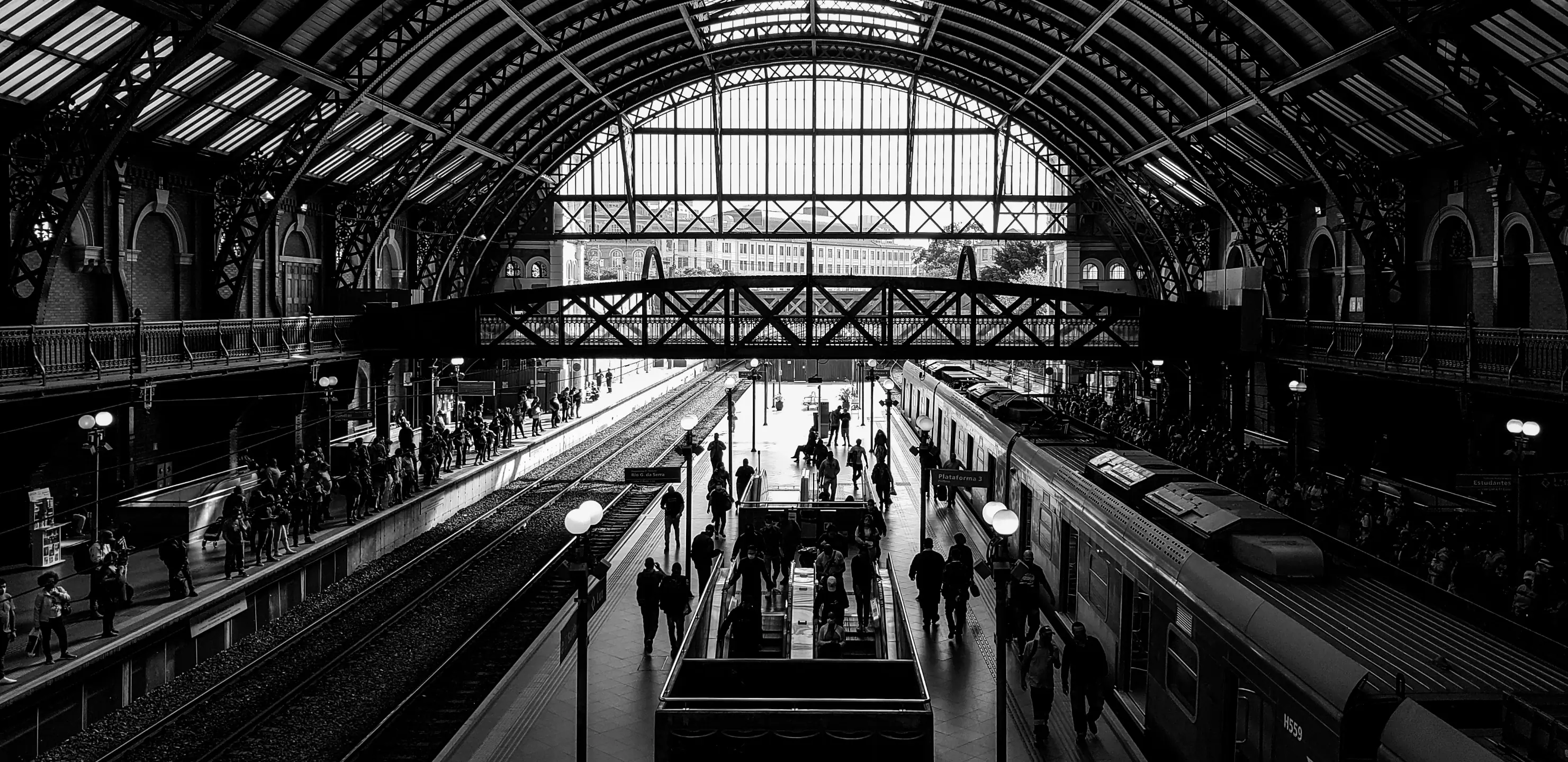 Interior de estação ferroviária com plataformas, estrutura metálica e circulação de pessoas