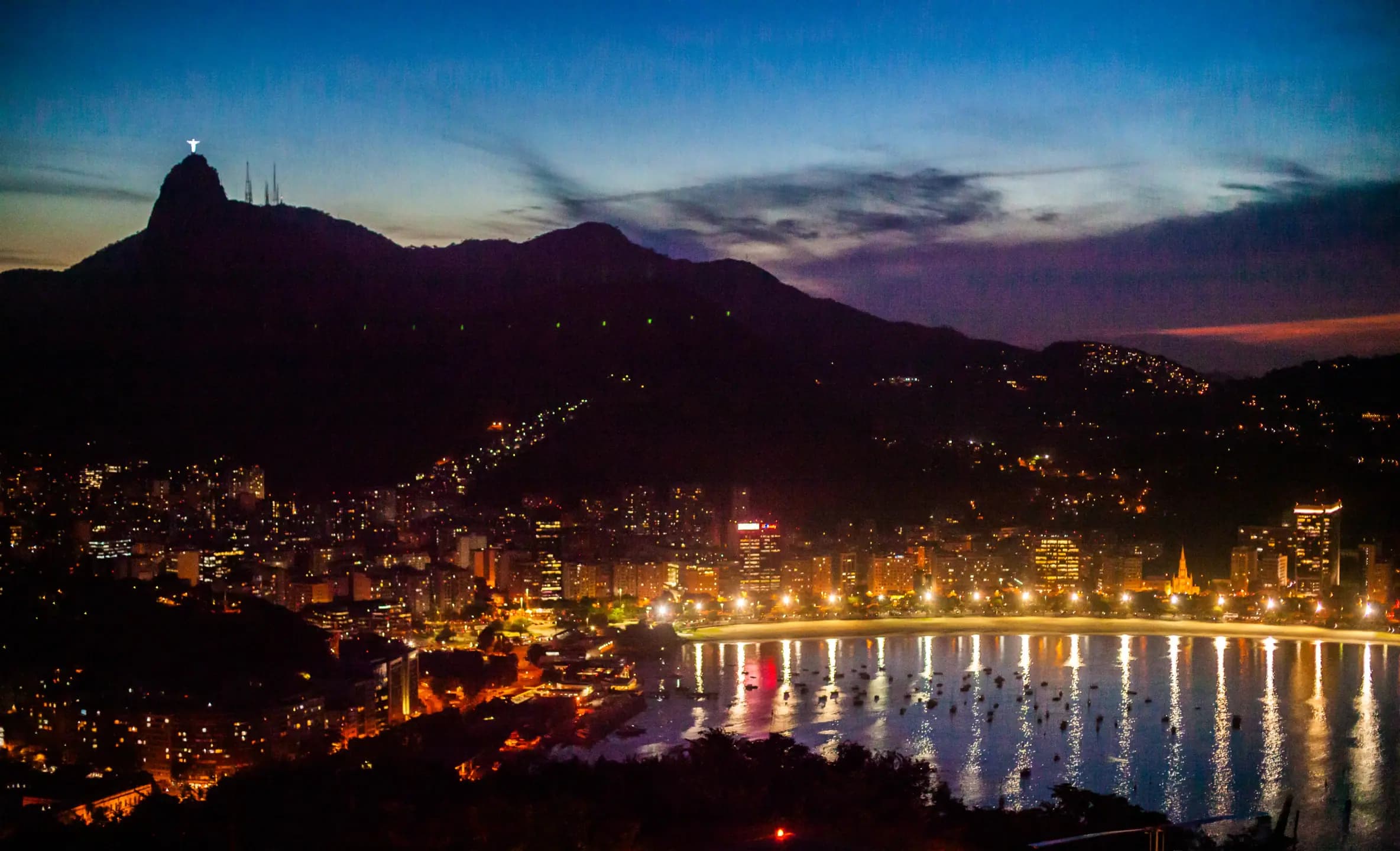 Baía com barcos, reflexos na água e morros ao fundo no anoitecer